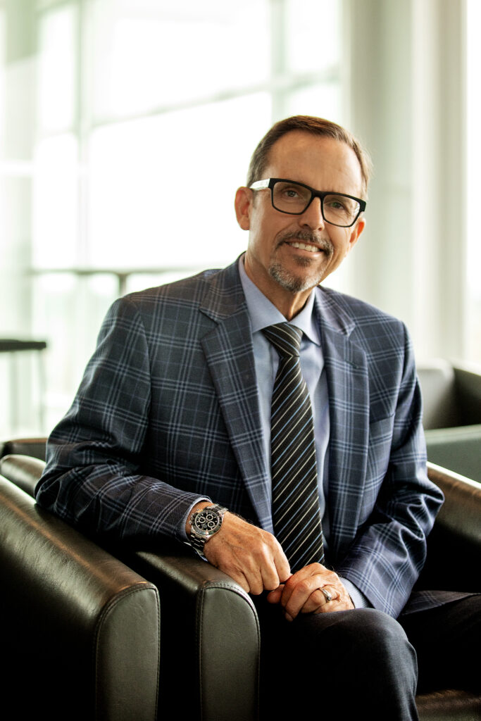 A person wearing a suit and glasses sits in a chair and smiles.