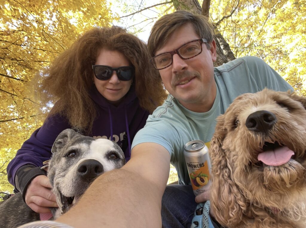 Two adults and two dogs take a selfie in front of a tree with yellow leaves. 