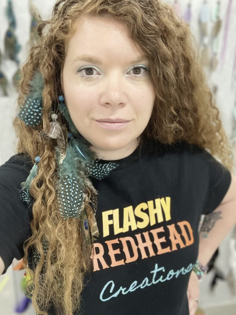 A person with long hair and feather hair extensions takes a selfie. Their T-shirt reads "Flashy Redhead Creations."
