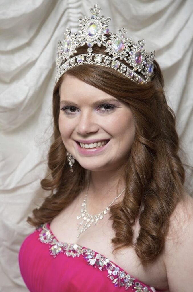 A person with long hair, wearing a sparkly crown, earrings, necklace and a pink dress. 