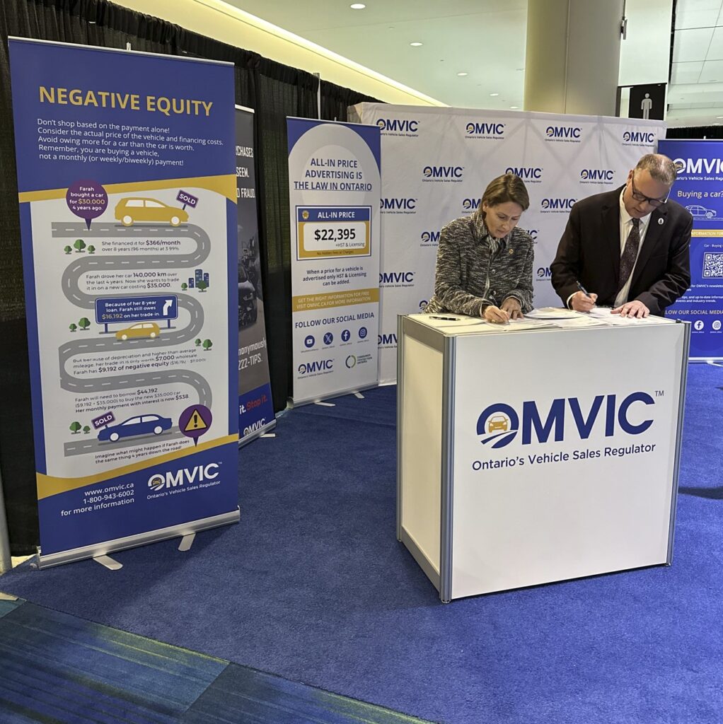Two people stand at a podium and sign a document in front of backdrops that read "OMVIC."
