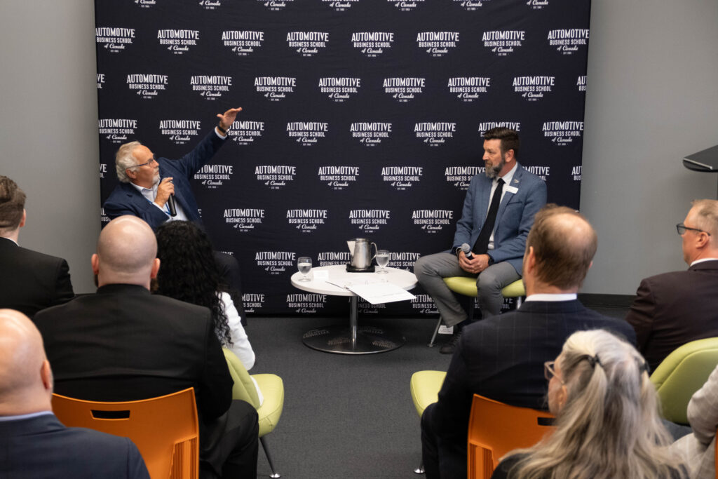 Two people sit and talk at the front of a room in front of a crowd of people. A backdrop reads: Automotive Business School of Canada.