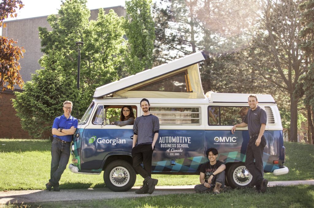 Five people pose inside or in front of a Volkswagen camper van that's parked outside in front of a bank of trees. 