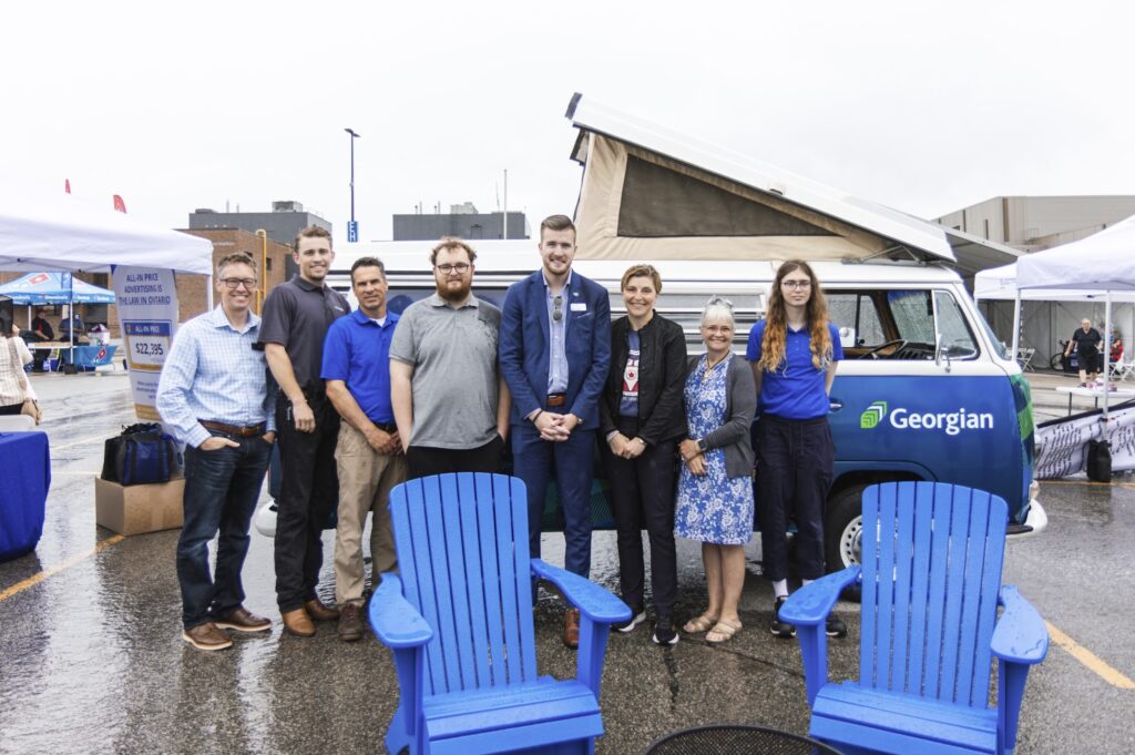 Eight people stand next to each other in front of a Volkswagen camper van that's parked in outside in a parking lot. Two blue Muskoka chairs and a firepit sit in front of the people.