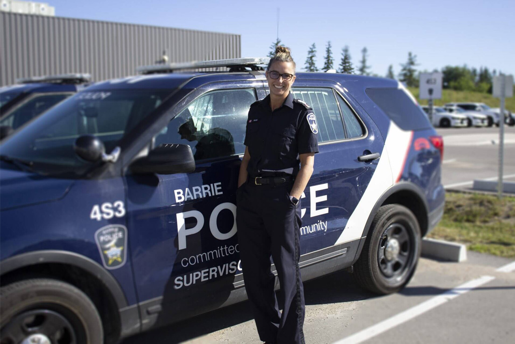 A person smiling, wearing glasses, with blonde hair tied up in a bun, wearing a Barrie Police Service uniform (navy collared shirt, black belt, navy trousers), standing next to a branded police SUV (Ford Explorer)