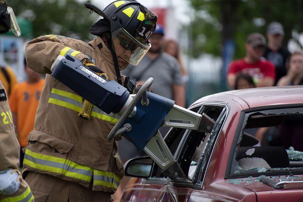 A firefighter from Barrie Fire and Emergency Services in uniform (tan suit and pants with yellow reflectors, sunglasses, and a black helmet with face shield) using an extrication cutter tool to open a car door after a car accident, as part of a demontration
