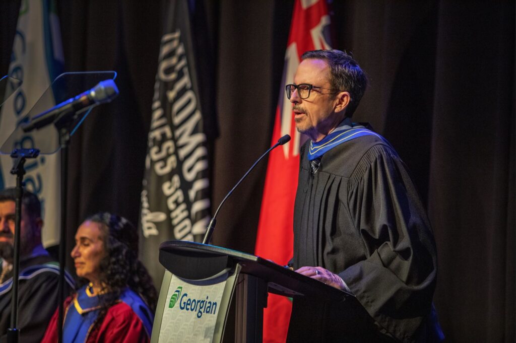 A person wearing a long black robe and glasses speaks at a podium.