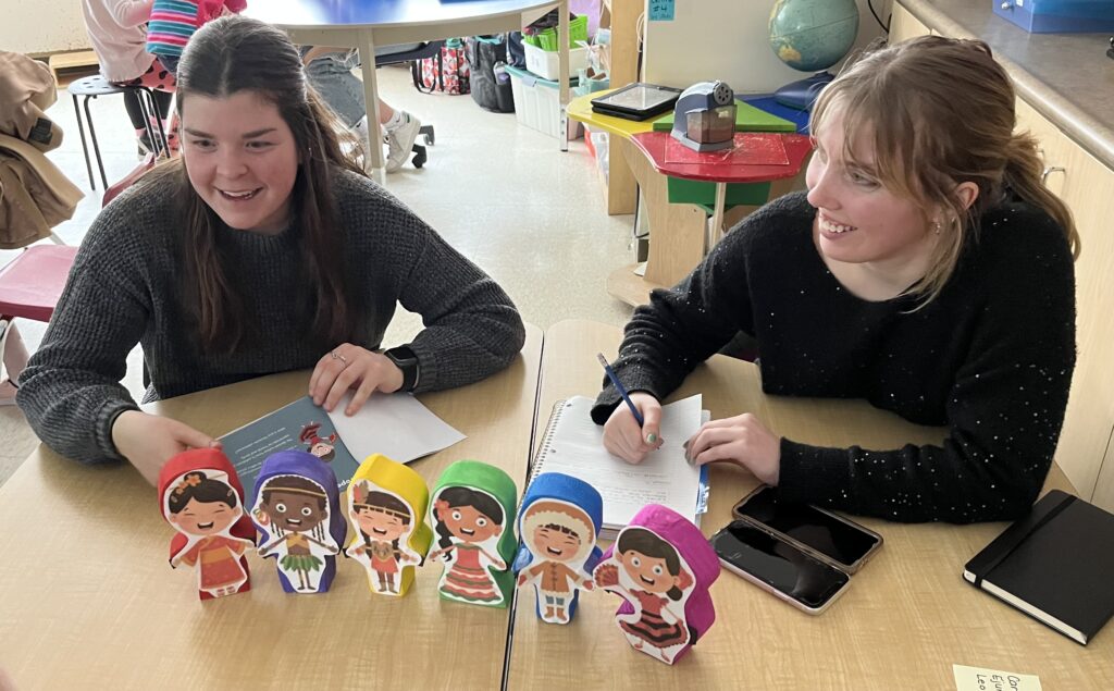 Two people wearing black sweaters sitting at a table in a classroom with a set of wooden dolls in different colours (red, purple, yellow, green, blue and pink).