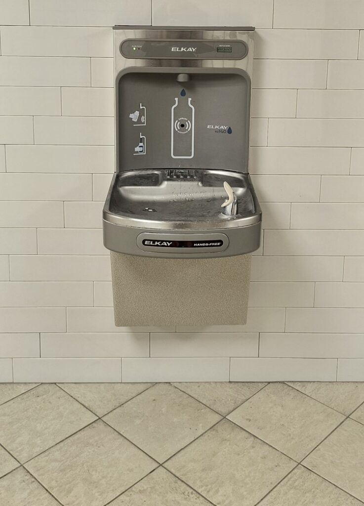 A metal water fountain and water dispenser is attached to a wall in a hallway. 