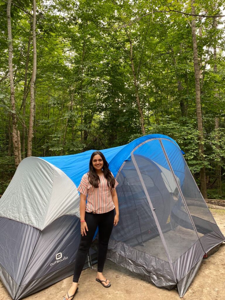 A person dressed casually stands outside in the woods in front of a blue-and-grey tent.