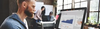Big Data Analyst with brown, slicked back hair and a beard using a Mac computer to analyze data reports in an office setting