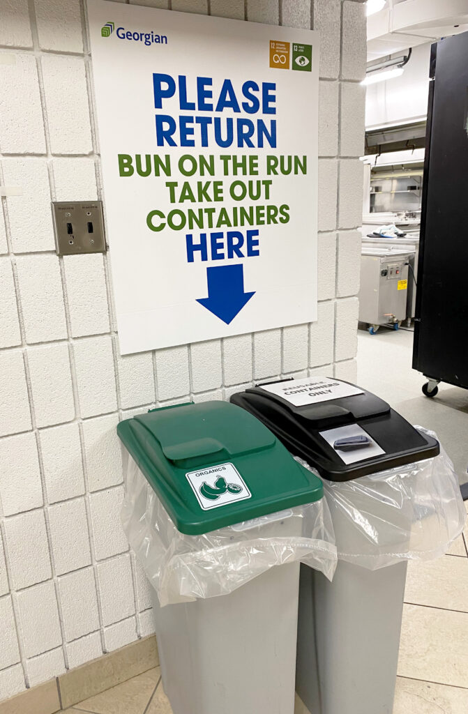 Plastic bins for organic waste and recyclables sit in a hallway next to a kitchen. Sign above reads: Return Bun on the Run takeout containers here.