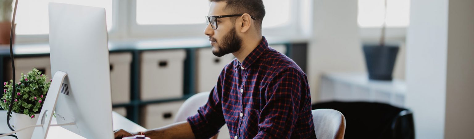 Student with dark hair, glasses and a yellow shirt sitting at a desk analyzing a spreadsheet on the computer and taking notes