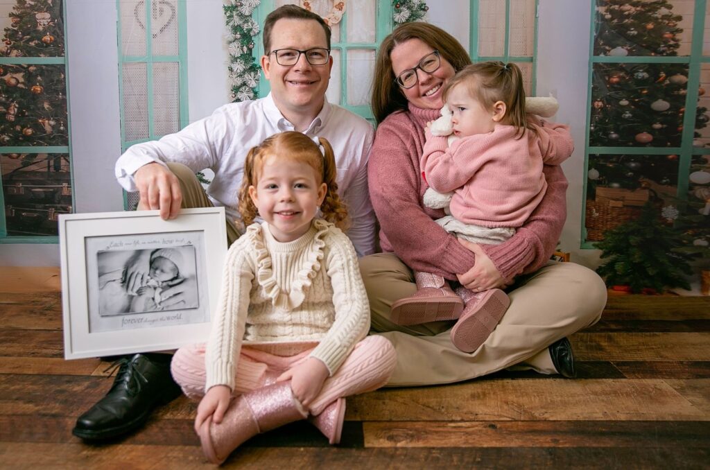Two adults and two children sit on the floor together, while one of the adults holds a framed photo of a baby.
