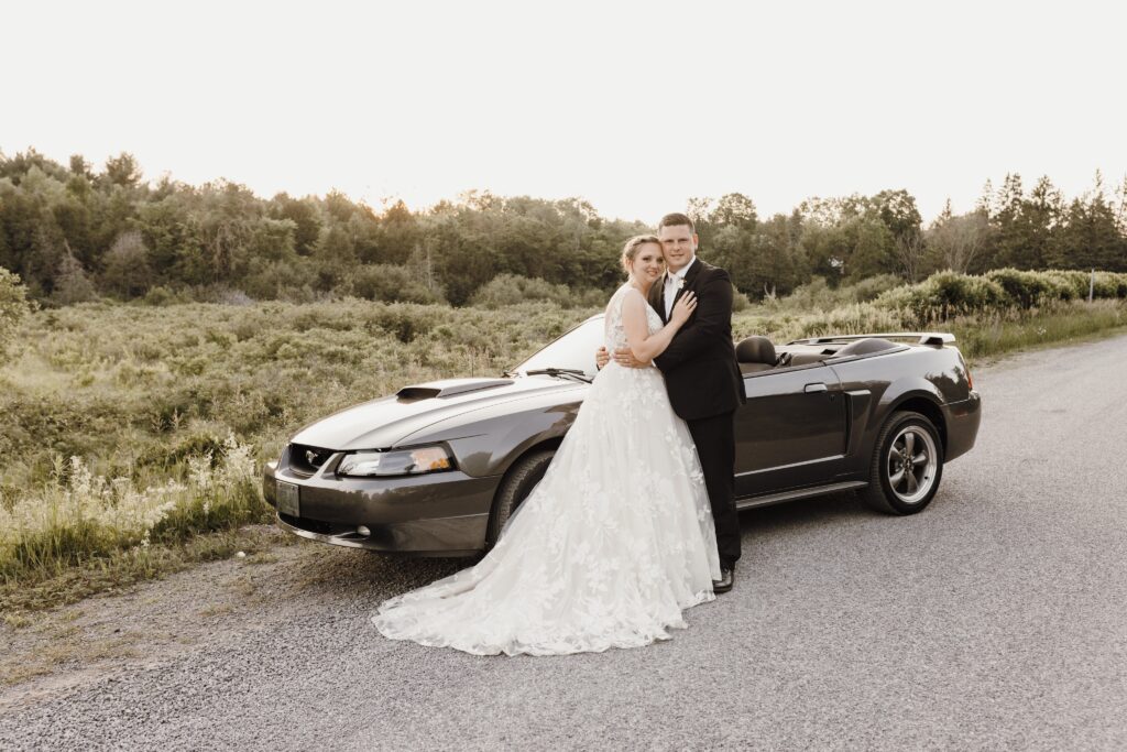 A bride and groom stand next to a car parked at the side of a road near a forest.