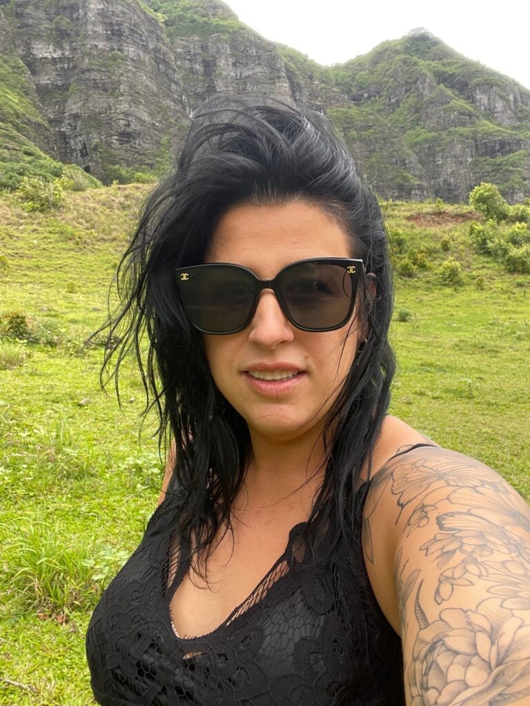 A person with long black hair, sunglasses and black tank top takes a selfie in front of a lush green, mountainous landscape.