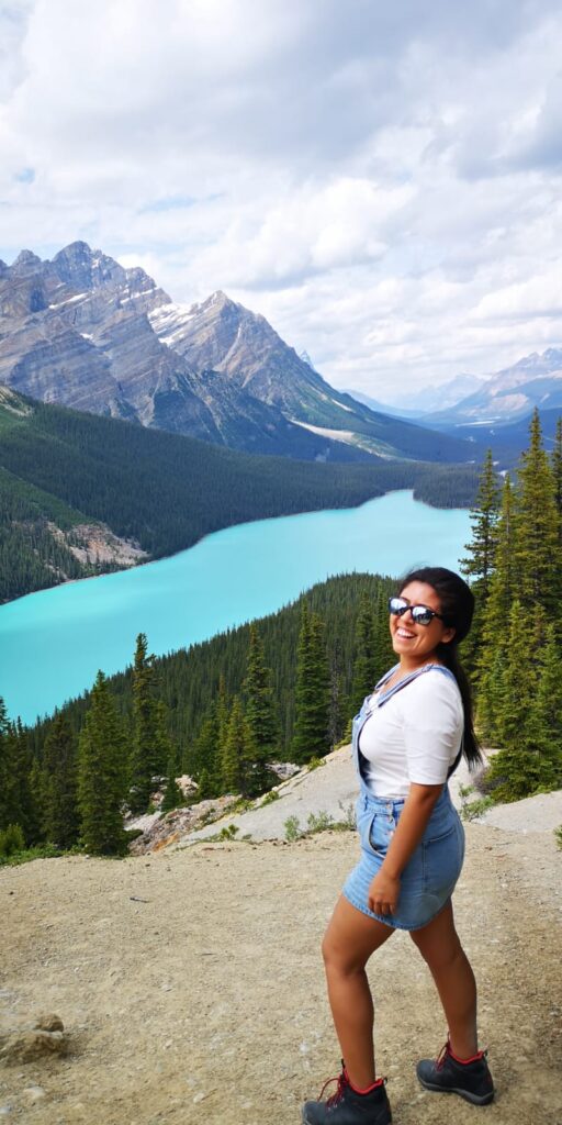 A person stands outside on a rock cliff overlooking a mountain range and bright blue lake.