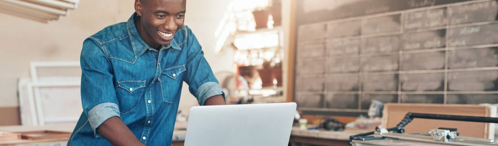 Entrepreneur wearing a denim collared shirt and smiling while standing at a counter using a laptop in a workshop setting
