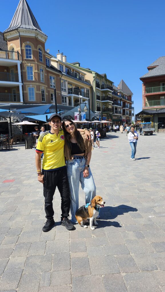 Two people and a dog stand outside with buildings and storefronts behind them.