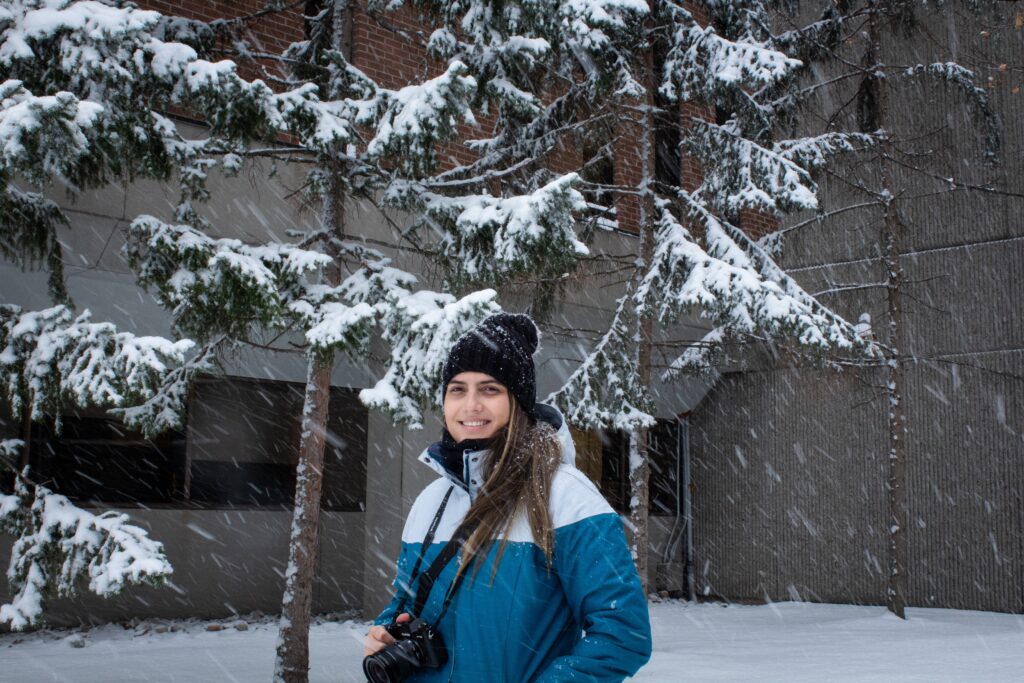 A person wearing a winter jacket and toque and holding a camera stands outside in the snow. 