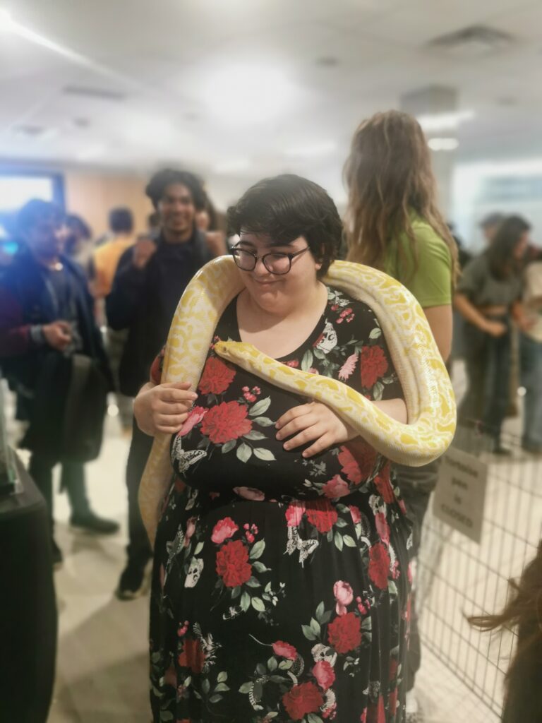 A person stands in a room surrounded by people with a large snake wrapped around their shoulders.