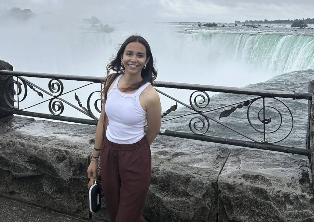 A person smiles while standing next to a railing with Niagara Falls behind them. 