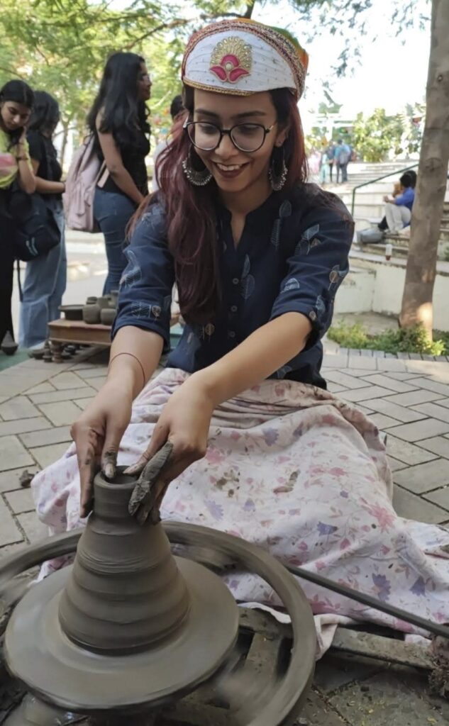 A person sits on the ground outside and uses their hands to shape pottery on a pottery wheel. 