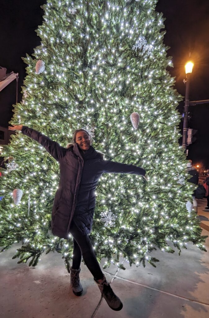 A person stands with arms outstretched in front of an outdoor tree covered in white lights. 