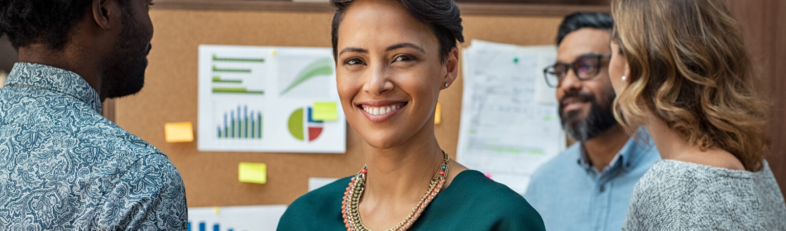 An accounting professional wearing an emerald green blazer, beaded necklace with dark pinned-back hair, holding a tablet while standing in front on a bulletin board with charts and statements pinned to it