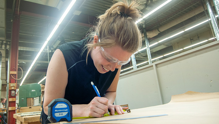 Cabinetmaking student wearing safety glasses while using a pencil and ruler to add markings to a drawing inside a work shop