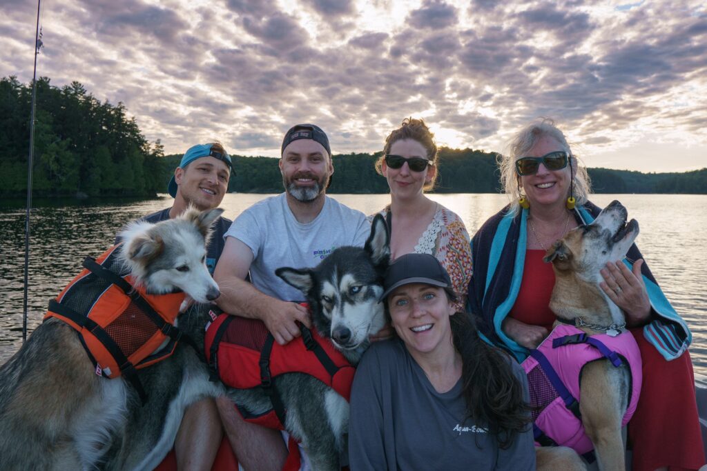 Five adults and three large dogs wearing life jackets sit together next to a lake near sunset.
