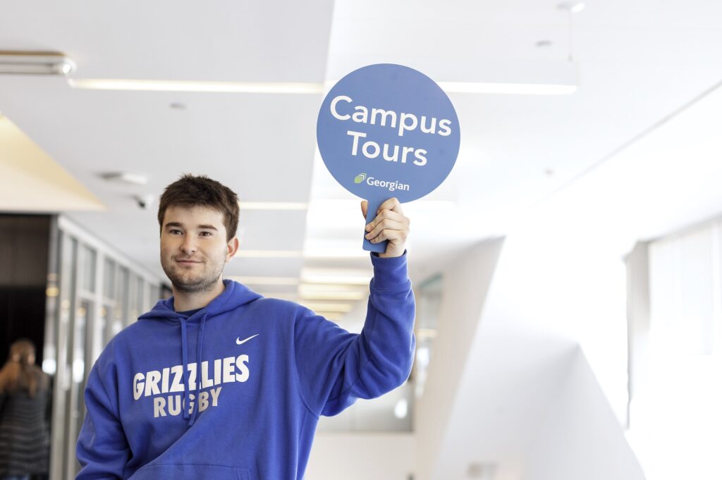 A person wearing a sweatshirt reading "Grizzlies Rugby" holds a sign reading "Campus Tours."