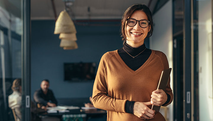 Person standing in an office setting while holding a tablet and wearing dark framed classes, a black turtle neck shirt with a rust coloured shirt over top