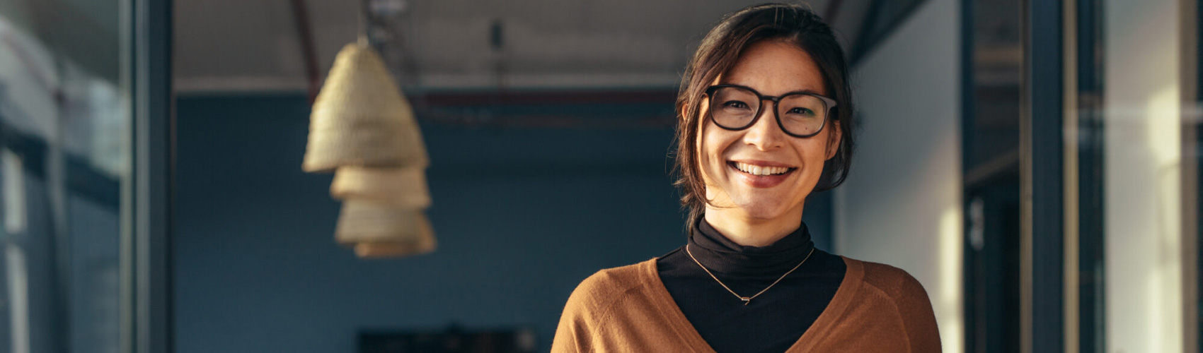 Person standing in an office setting while holding a tablet and wearing dark framed classes, a black turtle neck shirt with a rust coloured shirt over top