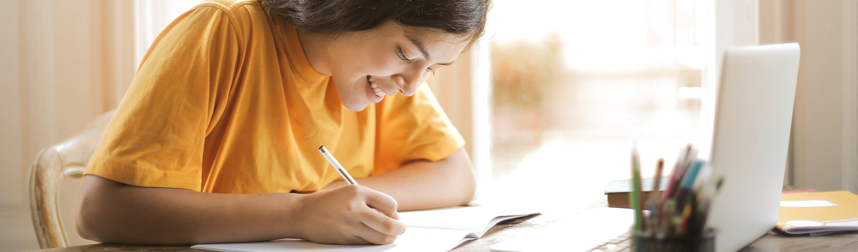 A youth with brown hair and a yellow t-shirt smiling while writing in a notebook at a desk