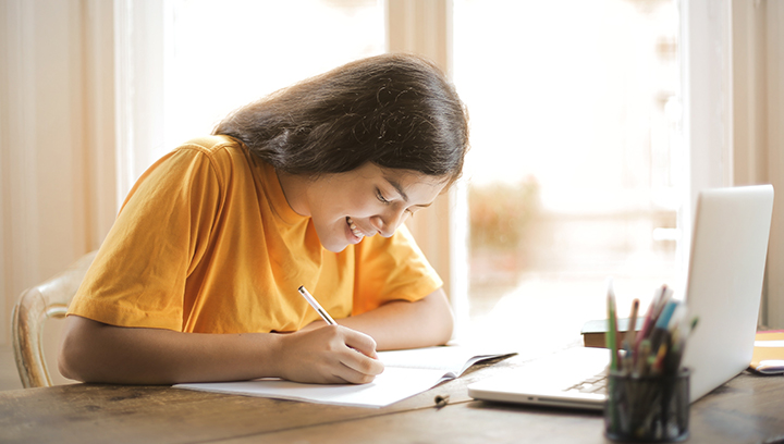 A youth with brown hair and a yellow t-shirt smiling while writing in a notebook at a desk