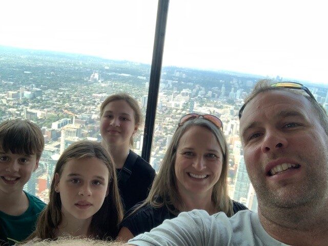 Two adults and three children take a selfie against a window with a sprawling view of a city.