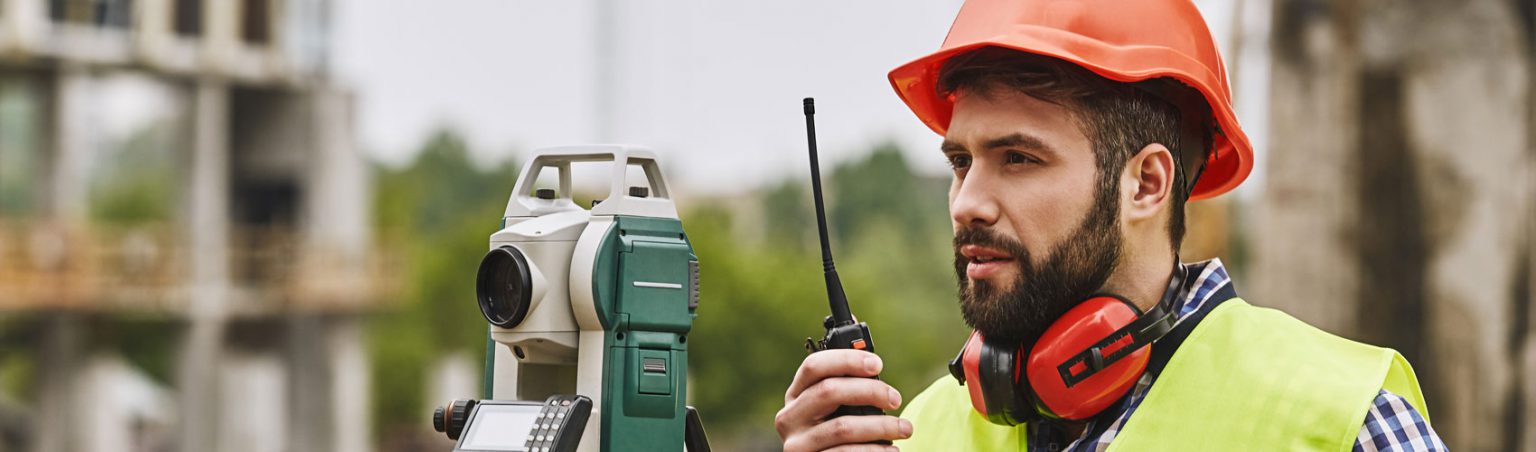 Civil Engineering Technician in a plaid shirt, safety vest and hard hat using a handheld transceiver and surveyor equipment