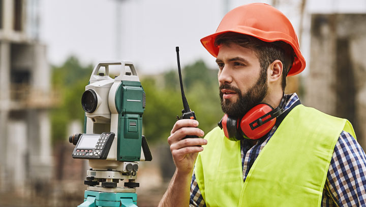 Civil Engineering Technician in a plaid shirt, safety vest and hard hat using a handheld transceiver and surveyor equipment