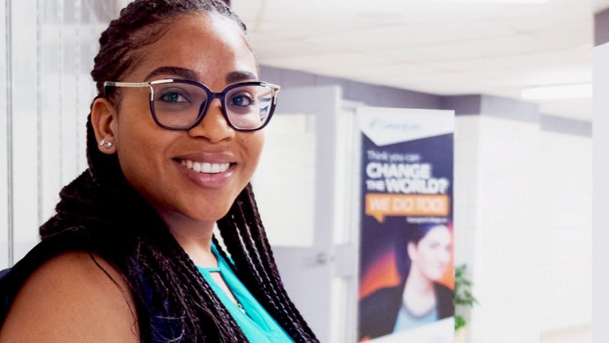 Female student wearing long braids and eyeglasses smiles at the camera in front of the offices of the Co-operative Education and Career Success office at the Barrie Campus.