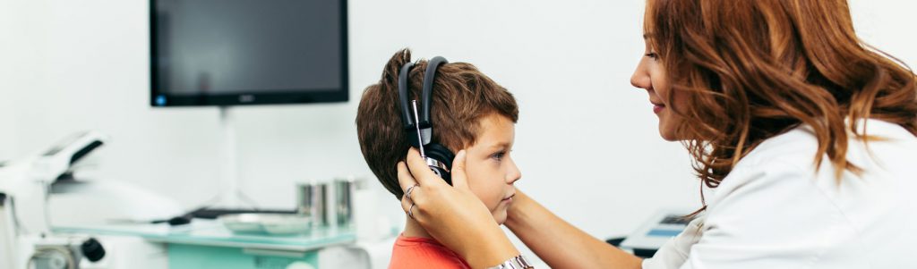 Communicative Disorders Assistant holding earphones over a young child's ears in a hearing clinic setting