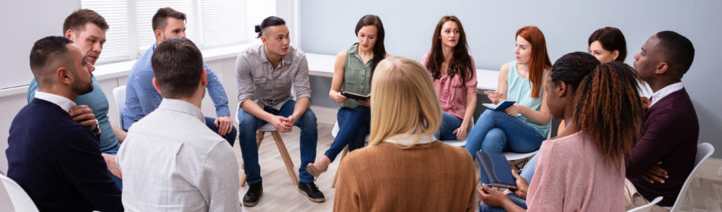A group of people sitting on chairs in a circle with pens and notebooks during a group counselling or intervention session