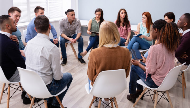 A group of people sitting on chairs in a circle with pens and notebooks during a group counselling or intervention session