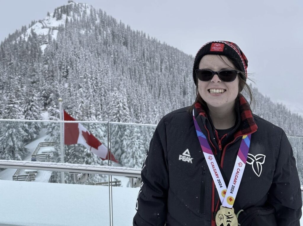 A person wearing a winter jacket, toque, sunglasses and a gold medal stands outside with a snowy mountain and Canada flag in the background.