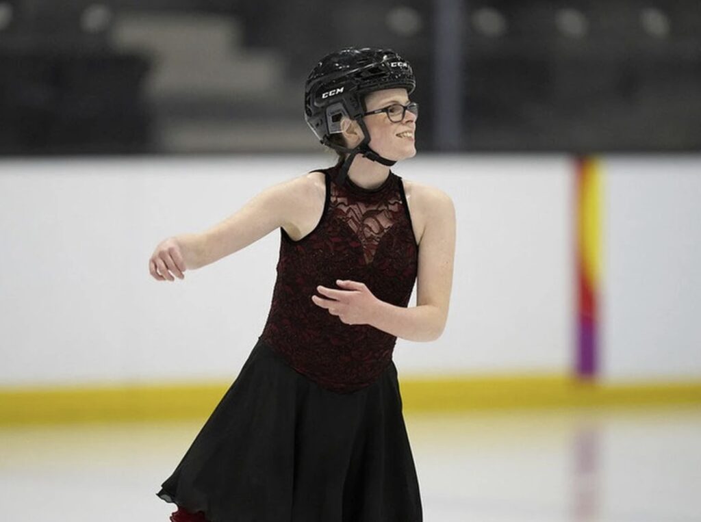A person wearing a dress and hockey helmet figure skates on an ice rink. 