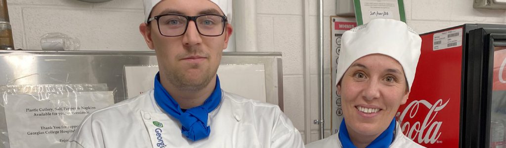 Two CICE students wearing chef's hats, jackets, blue neck scarves and name tags in the culinary lab at Georgian College