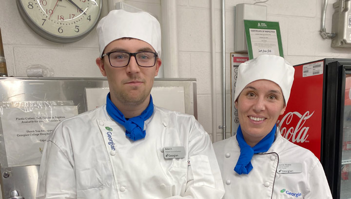 Two CICE students wearing chef's hats, jackets, blue neck scarves and name tags in the culinary lab at Georgian College