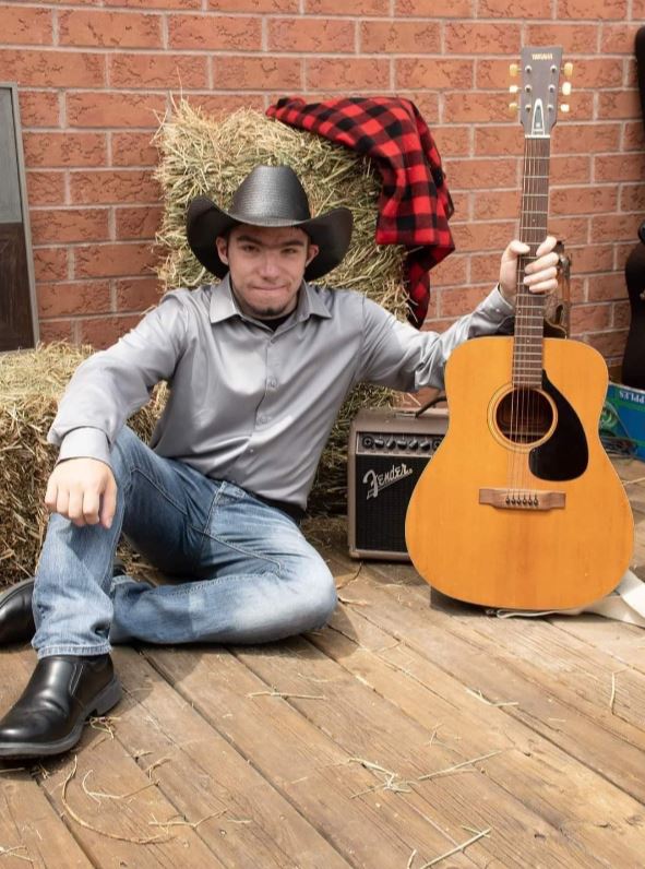 A person wearing a black cowboy hat, grey dress shirt, blue jeans, black shoes, and holding a guitar, sits on a deck in front of haybales.