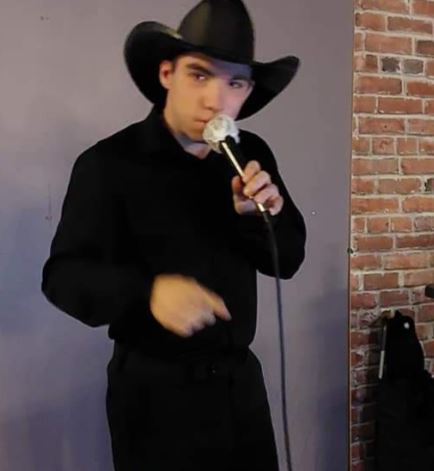 A person dressed all in black, including a cowboy hat, holds a microphone and sings on stage.