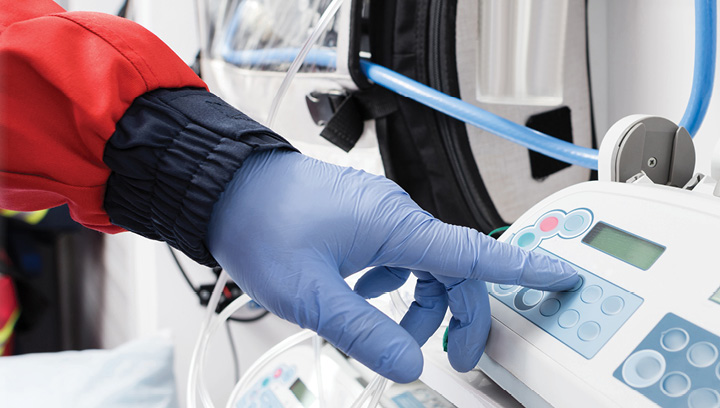 A community paramedic wearing blue latex gloves pushing a button on an IV pump preparing to administer in-home care to a community member